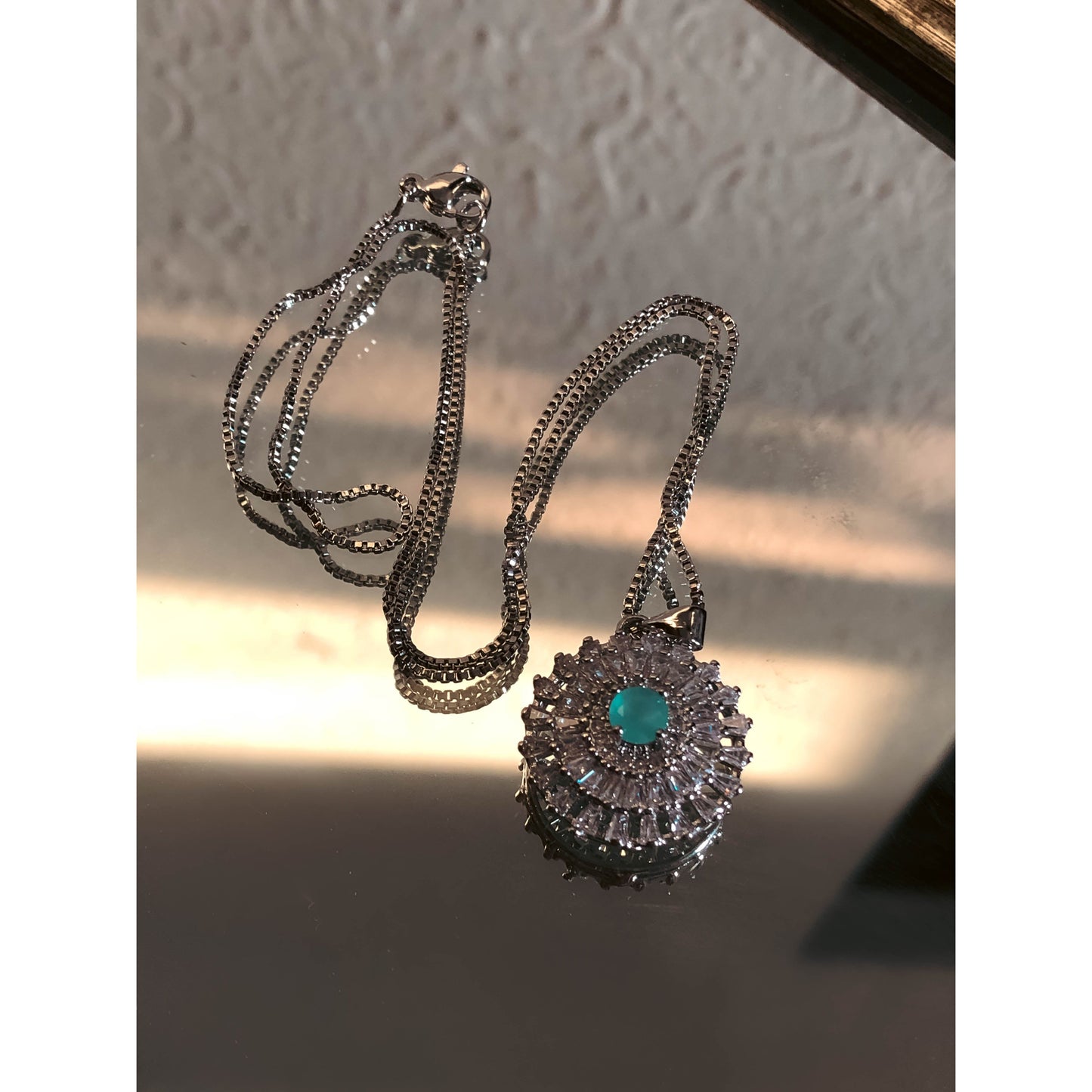 A close-up of a silver-tone necklace with a pendant featuring a large, clear, round stone surrounded by a halo of smaller clear sparkling stones, on a delicate chain.