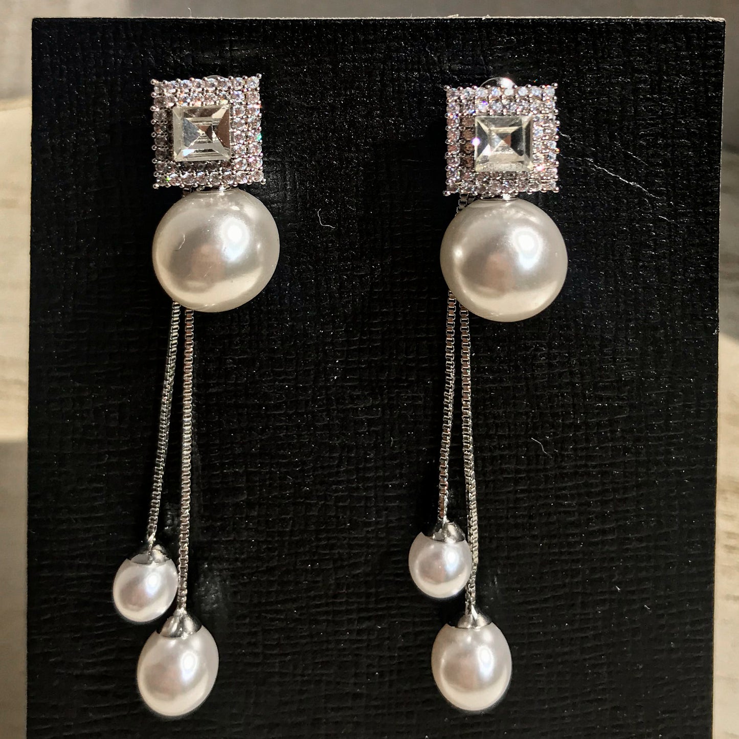 Two silver drop earrings, each with a square top of sparkling clear stones above a large white pearl, from which two chains with smaller white pearls dangle, displayed on a black textured surface.