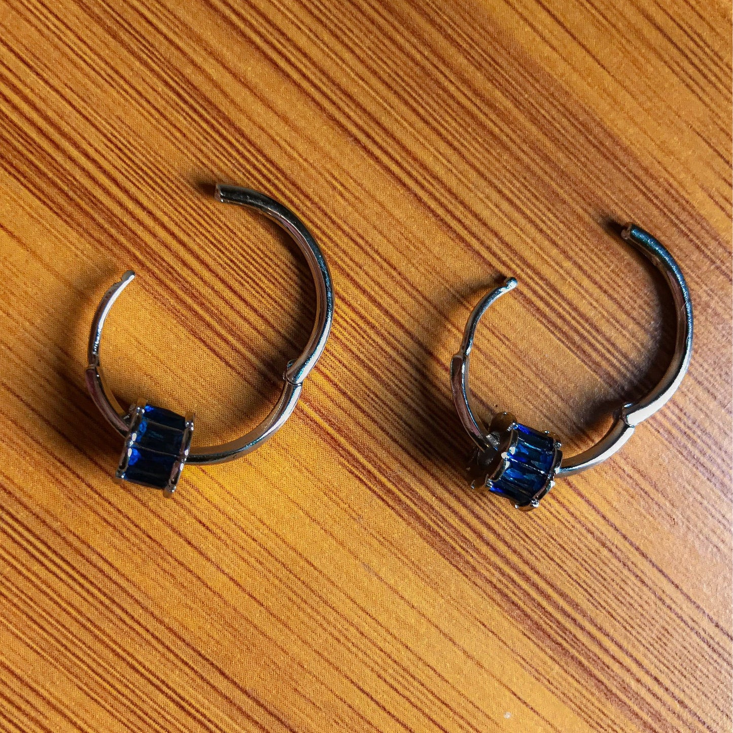 Two small silver hoop earrings, each with a cluster of rectangular blue stones on the front, resting on a wooden surface.