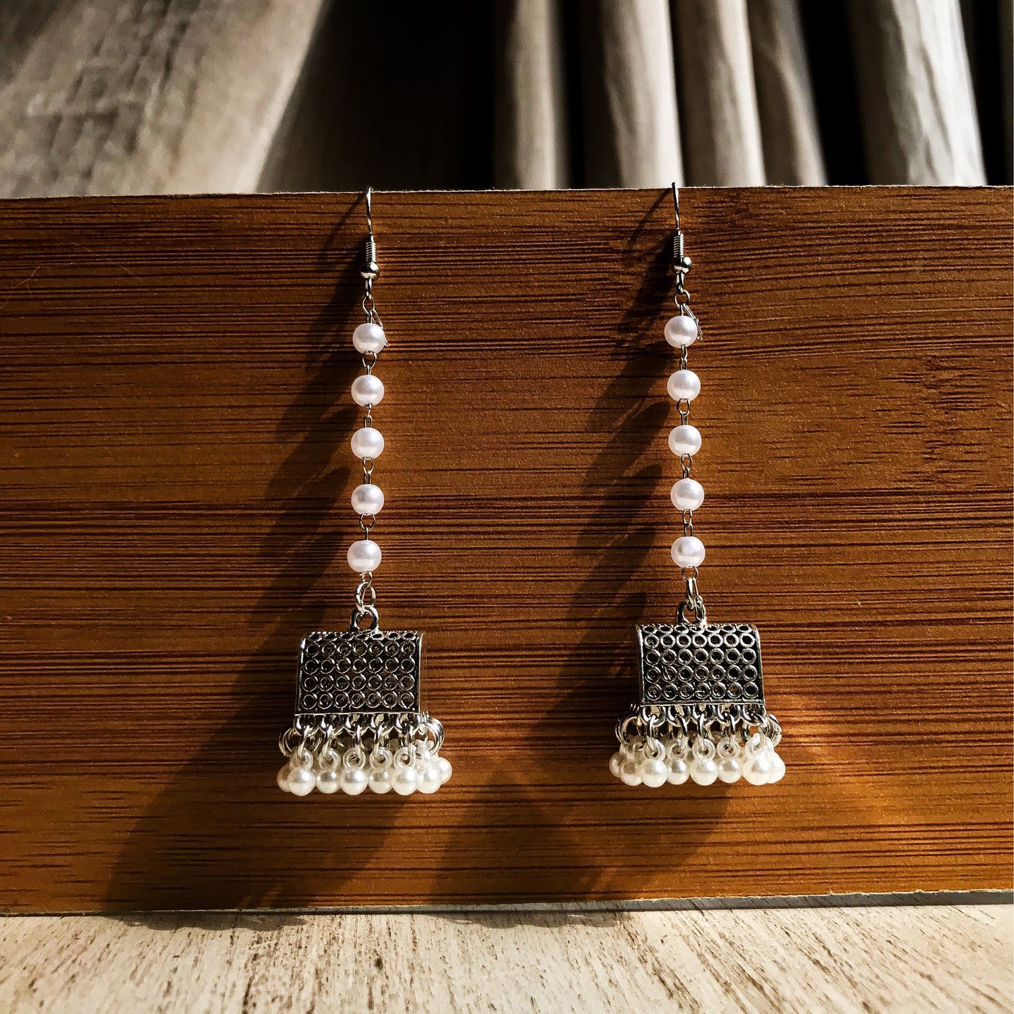 Two silver-tone dangle earrings with a textured geometric top, a chain of white pearls, and a fringe of small white pearls at the bottom, displayed against a wooden background.