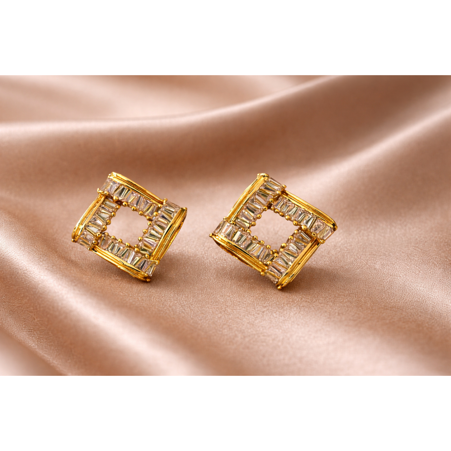 Pair of square frame gold-tone crystal stud earrings displayed on light brown satin fabric with soft folds, close-up jewellery product image