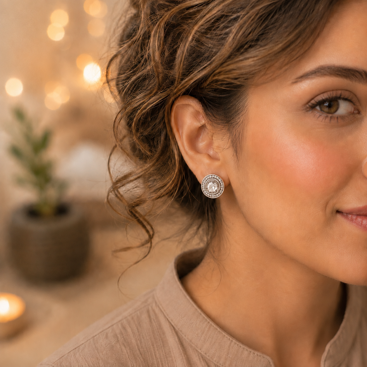 Close-up of a woman wearing silver halo stud earrings with a round clear crystal centre in a warm indoor setting with soft bokeh lighting.