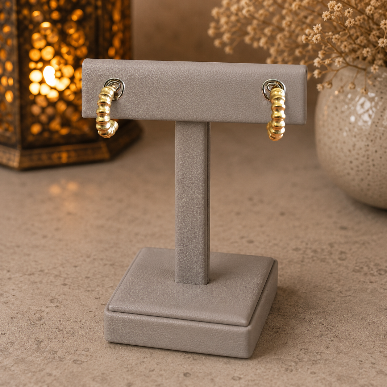 Textured gold mini hoop earrings displayed on a premium grey leather stand with a softly blurred lantern and ceramic vase background.