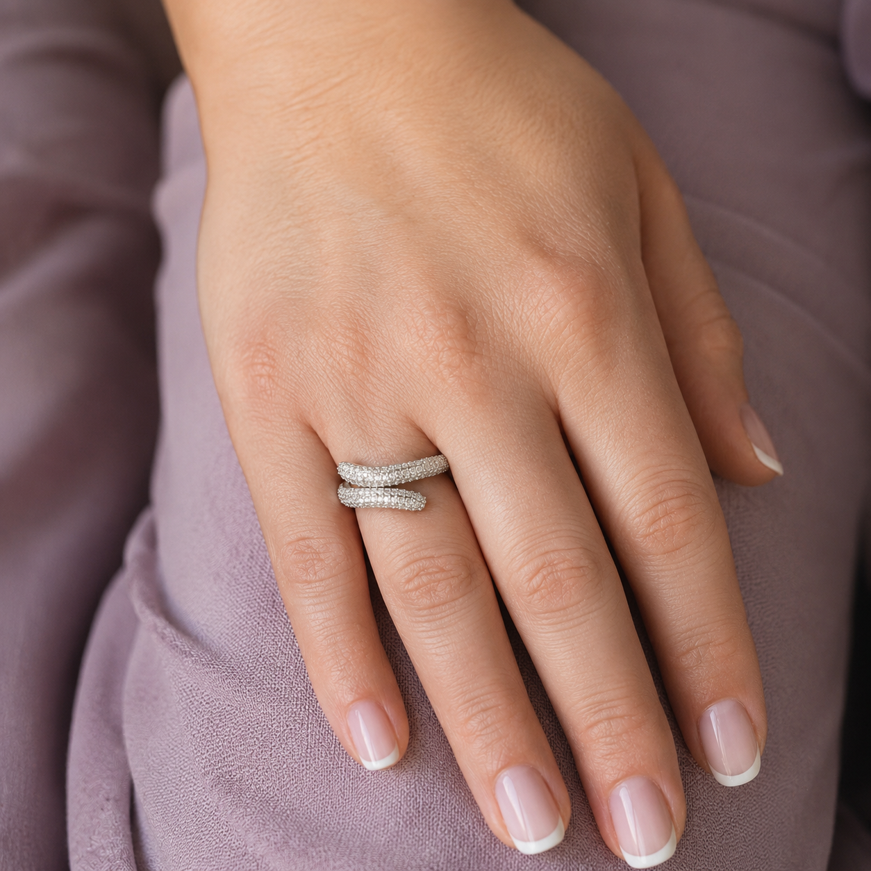 Woman wearing a silver twisted pavé wrap ring with sparkling micro-set stones on her hand, photographed in a luxury editorial style with a soft mauve background