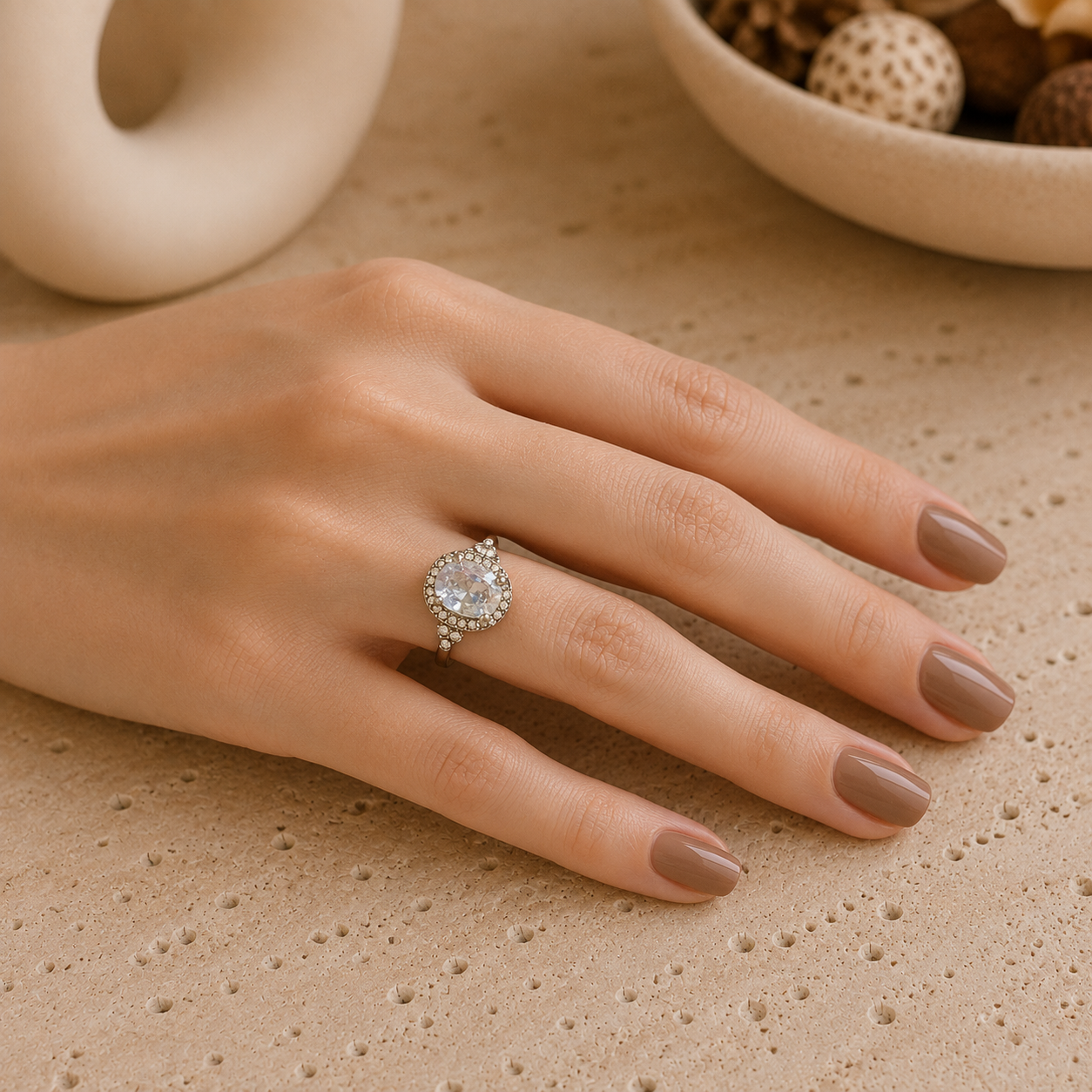 Silver-tone oval halo crystal ring worn on a feminine hand with taupe-brown manicure resting on a natural travertine surface.
