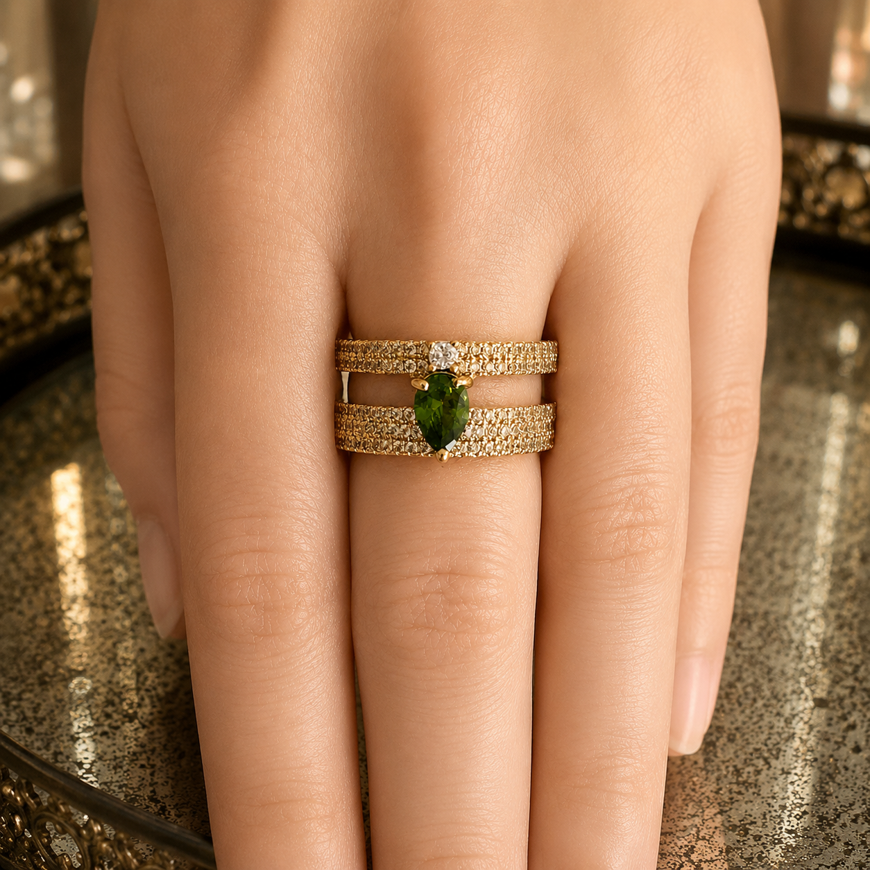 A close-up of a hand wearing a double gold ring set with pavé diamonds and a green marquise-cut gemstone at the center, resting on a reflective metallic surface.