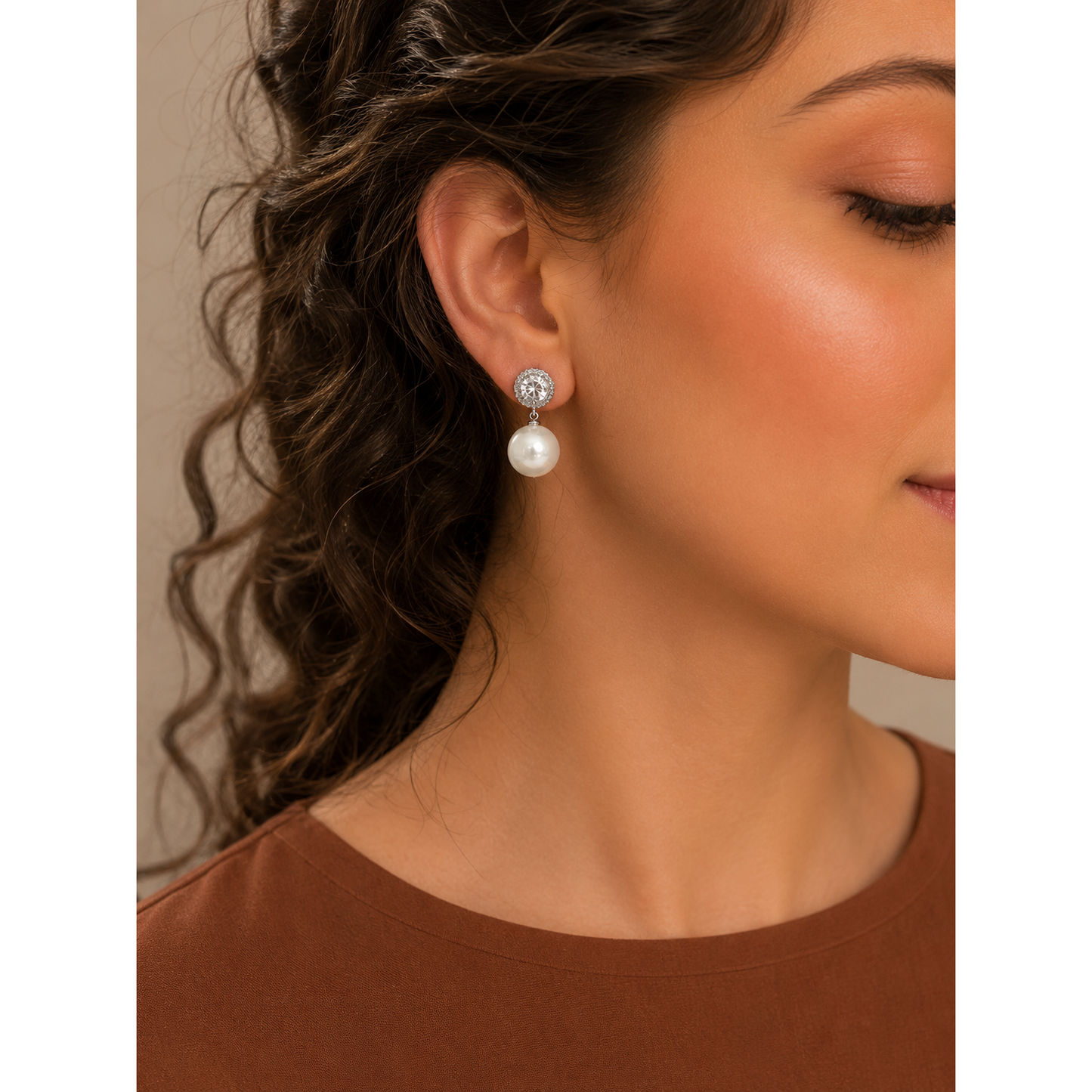Woman wearing silver crystal halo pearl drop earrings with soft brown curls and a copper-brown satin blouse in a studio portrait.