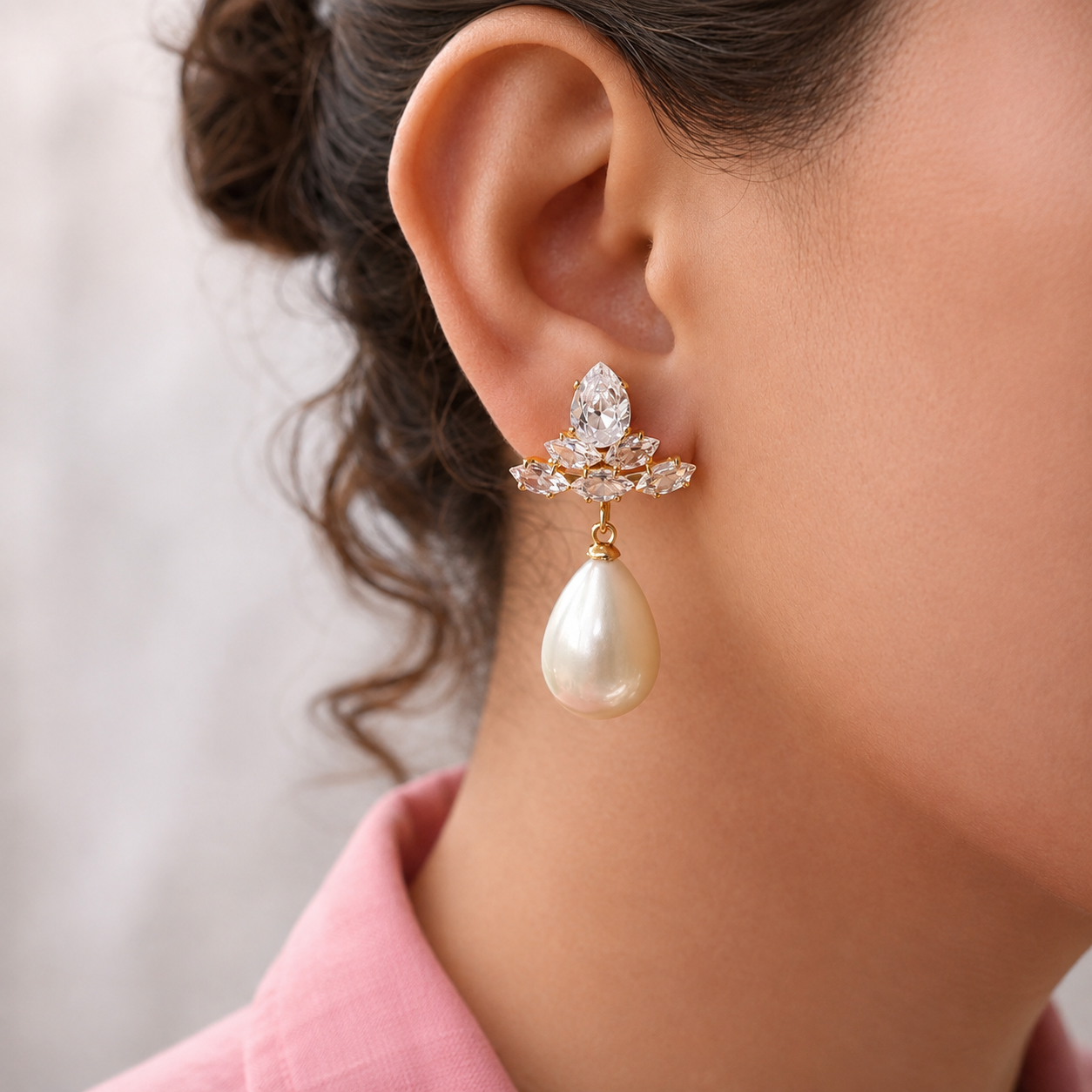 A close-up of a model's ear wearing a gold and crystal drop earring with a teardrop-shaped pearl pendant, paired with a pink collared shirt against a soft neutral background.