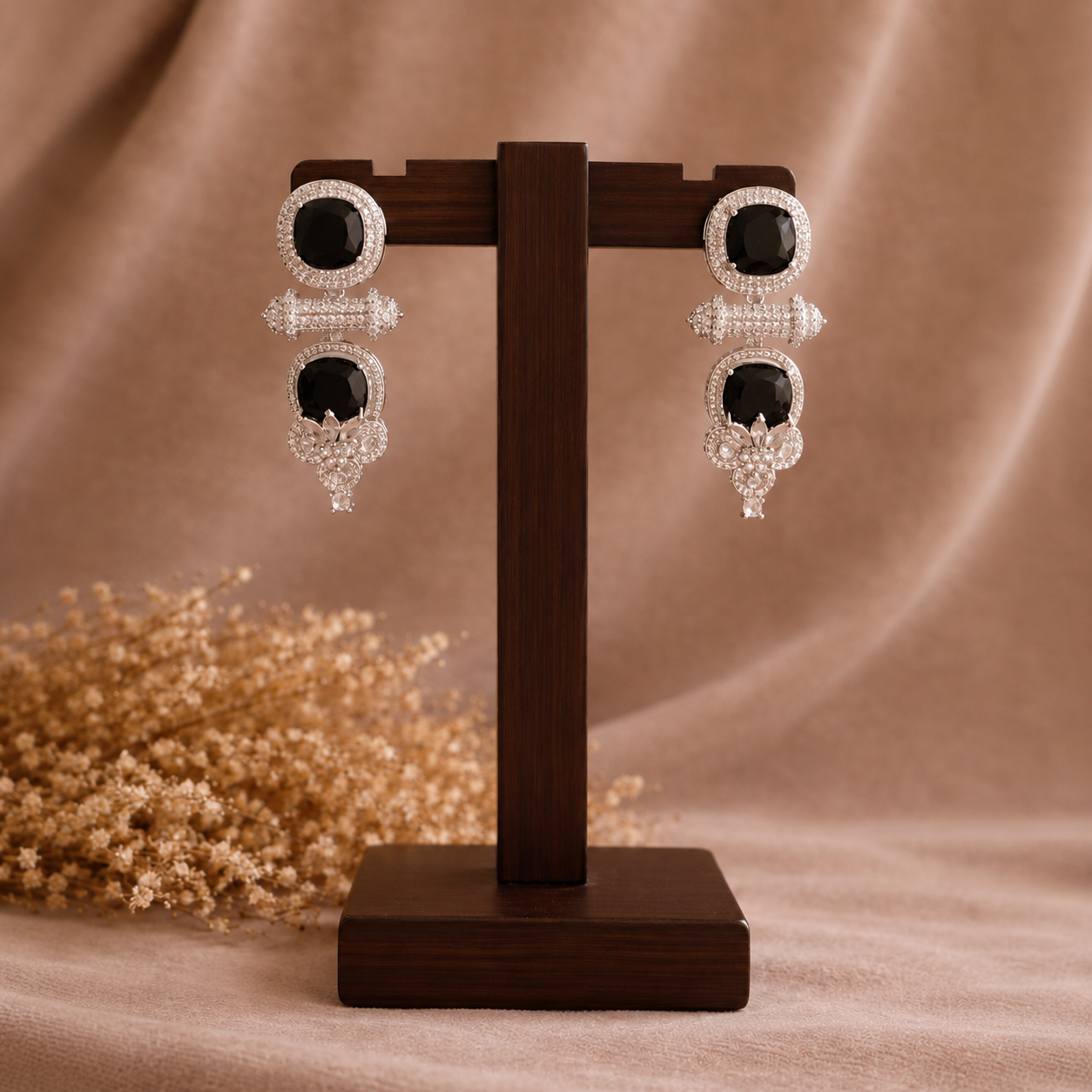 A pair of silver-toned drop earrings with large black oval stones and clear crystal detailing, displayed on a dark wooden stand against a warm beige fabric backdrop with dried flowers.
