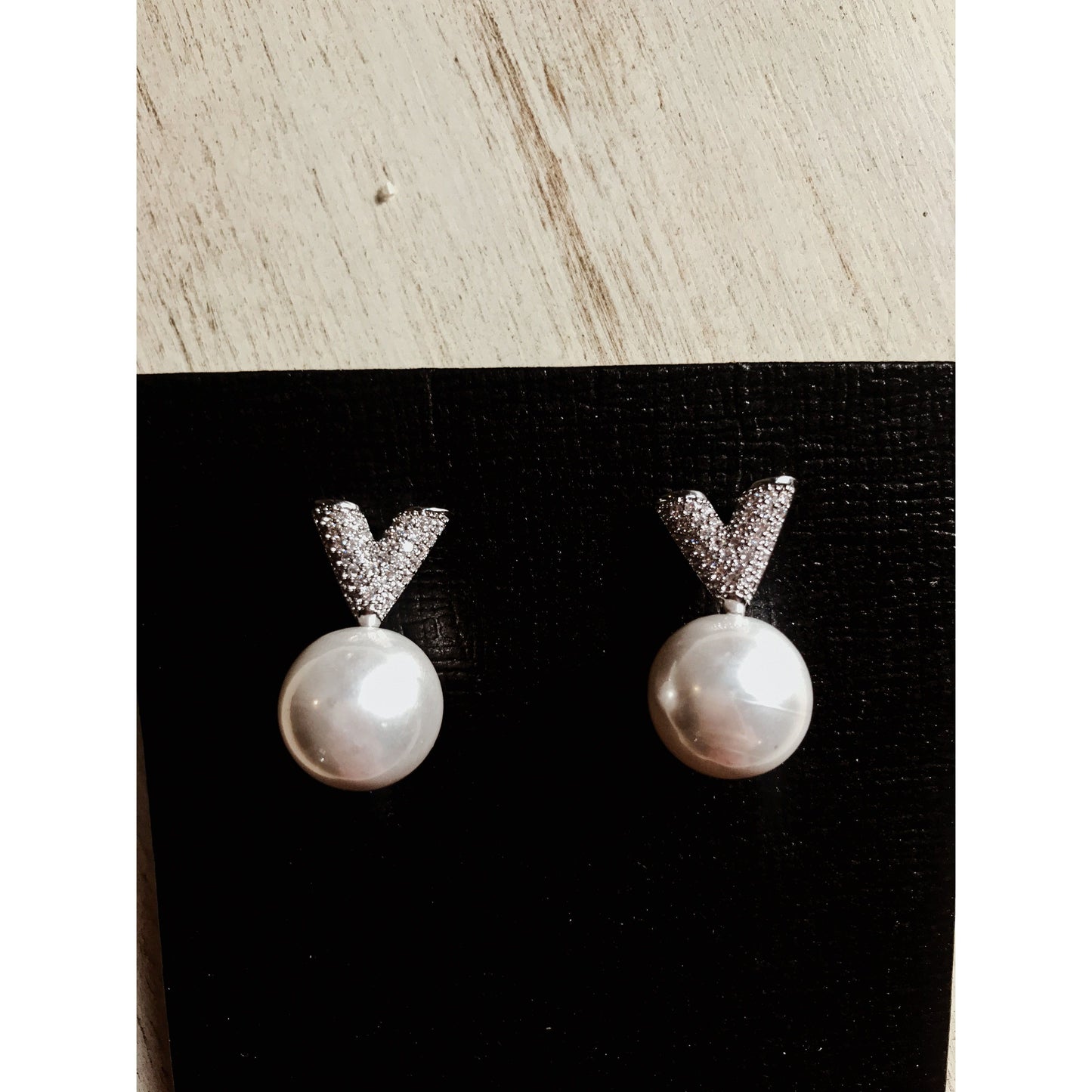 Two silver drop earrings, each featuring a white pearl hanging below a sparkling silver V-shape made of small clear stones, displayed on a textured black surface.