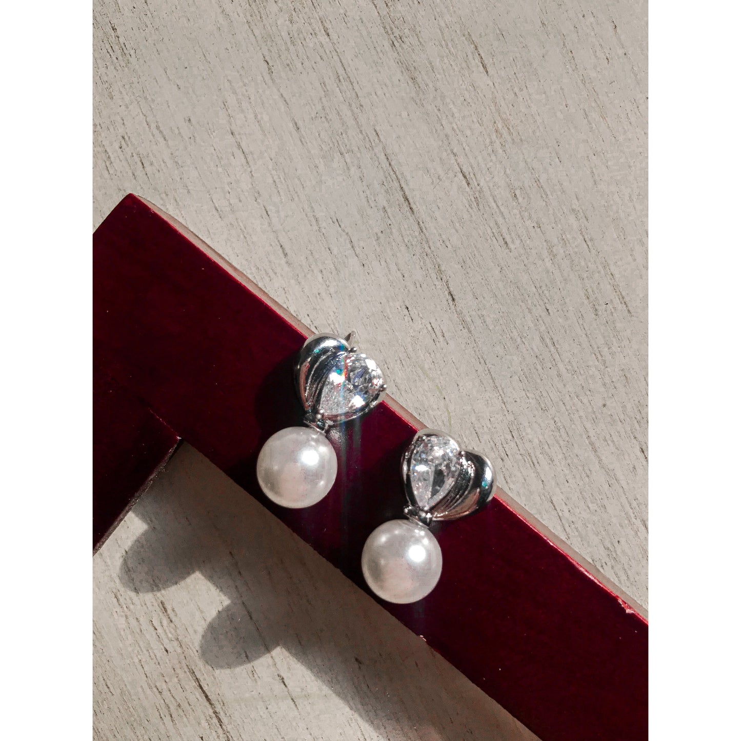 Two elegant silver drop earrings, each featuring a white pearl hanging below a silver heart with a clear, pear-shaped stone, displayed on a textured surface.