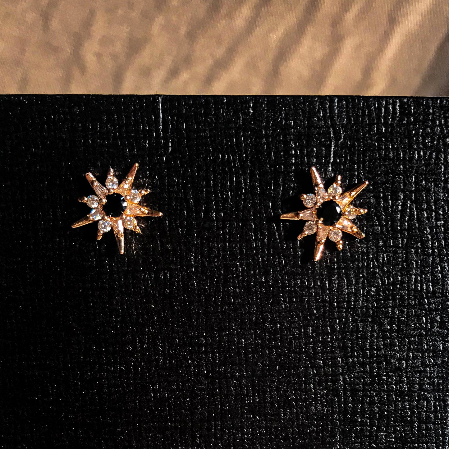 Two rose gold starburst earrings with a sparkling stone at the center, displayed on a black earring card.