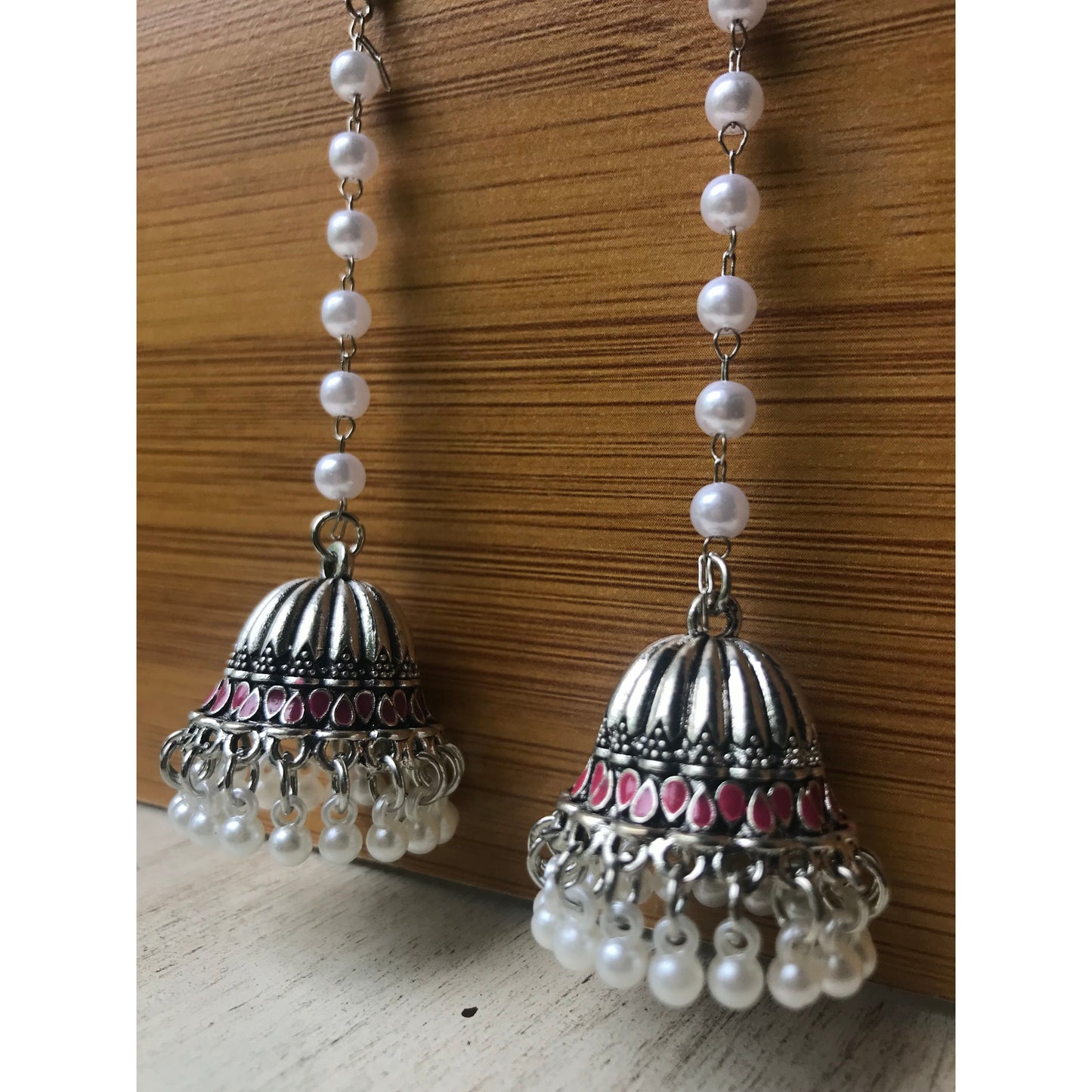 Pair of silver jhumka earrings with pink accents and pearl beaded chains hanging on a wooden backdrop against a white surface