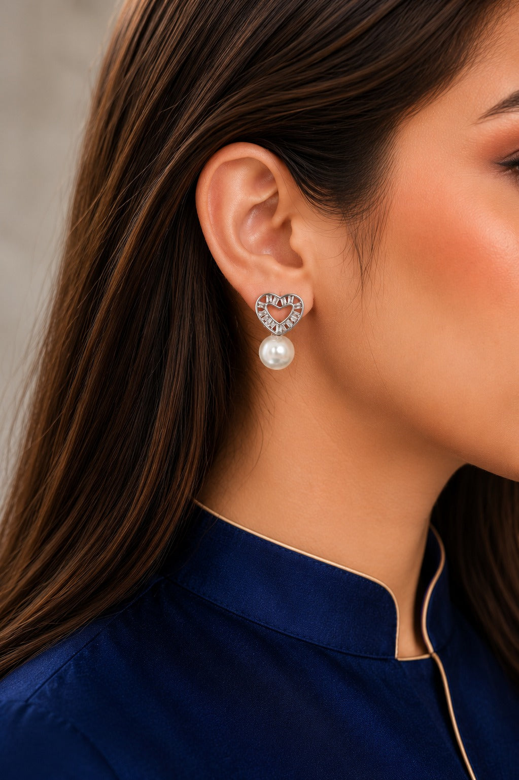 Silver heart-shaped cubic zirconia pearl stud earrings worn by model with navy satin shirt, featuring a lustrous pearl drop.
