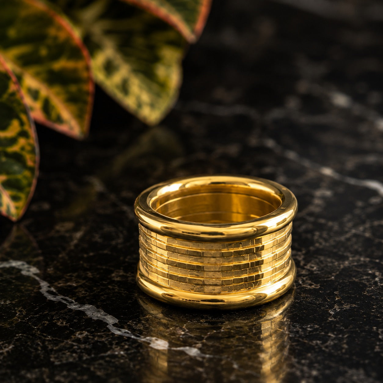 Wide polished stainless steel ring showcased on a black marble tabletop with decorative foliage.