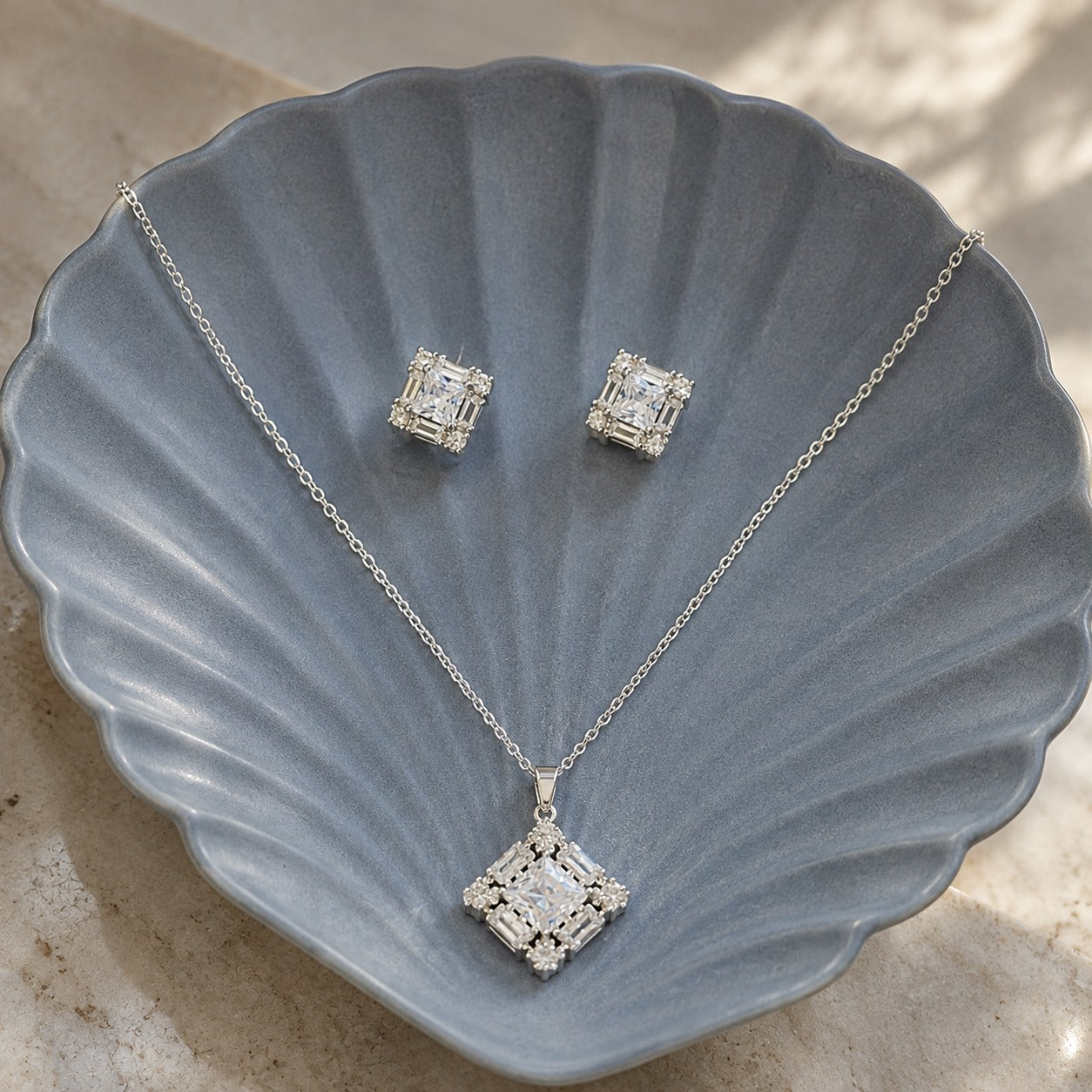 Facet Grid S925 silver earrings and pendant set with square clear stones and baguette-style zircon detailing displayed on a grey shell-shaped plate