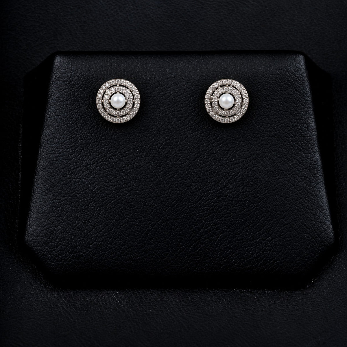 Pair of Pearl Axis Studs displayed on a black jewellery stand, featuring white pearl centres and double concentric zircon circles in silver-tone metal.