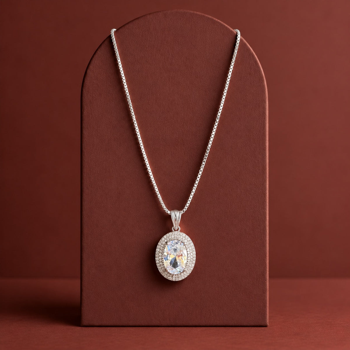 Silver-tone Aurelle Oval Zircon Pendant with a coordinating chain, showcasing its oval-cut crystal-clear centre stone and double zircon border on a premium terracotta jewellery display.