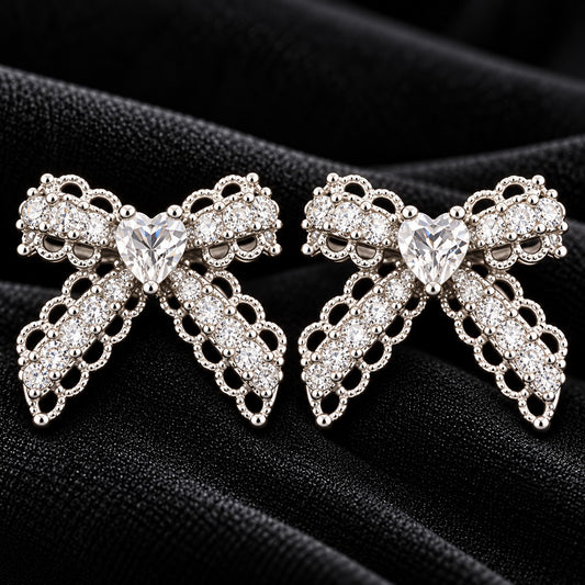 Close-up of Amorette Bow Studs featuring clear heart-shaped centre stones, clear zircon detailing, and polished silver-tone bow designs on black fabric.