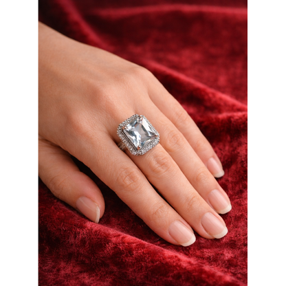 hand wearing an elegant cushion-cut cubic zirconia ring with a double halo of smaller stones resting on red velvet fabric, showcasing a luxury statement jewelry piece.