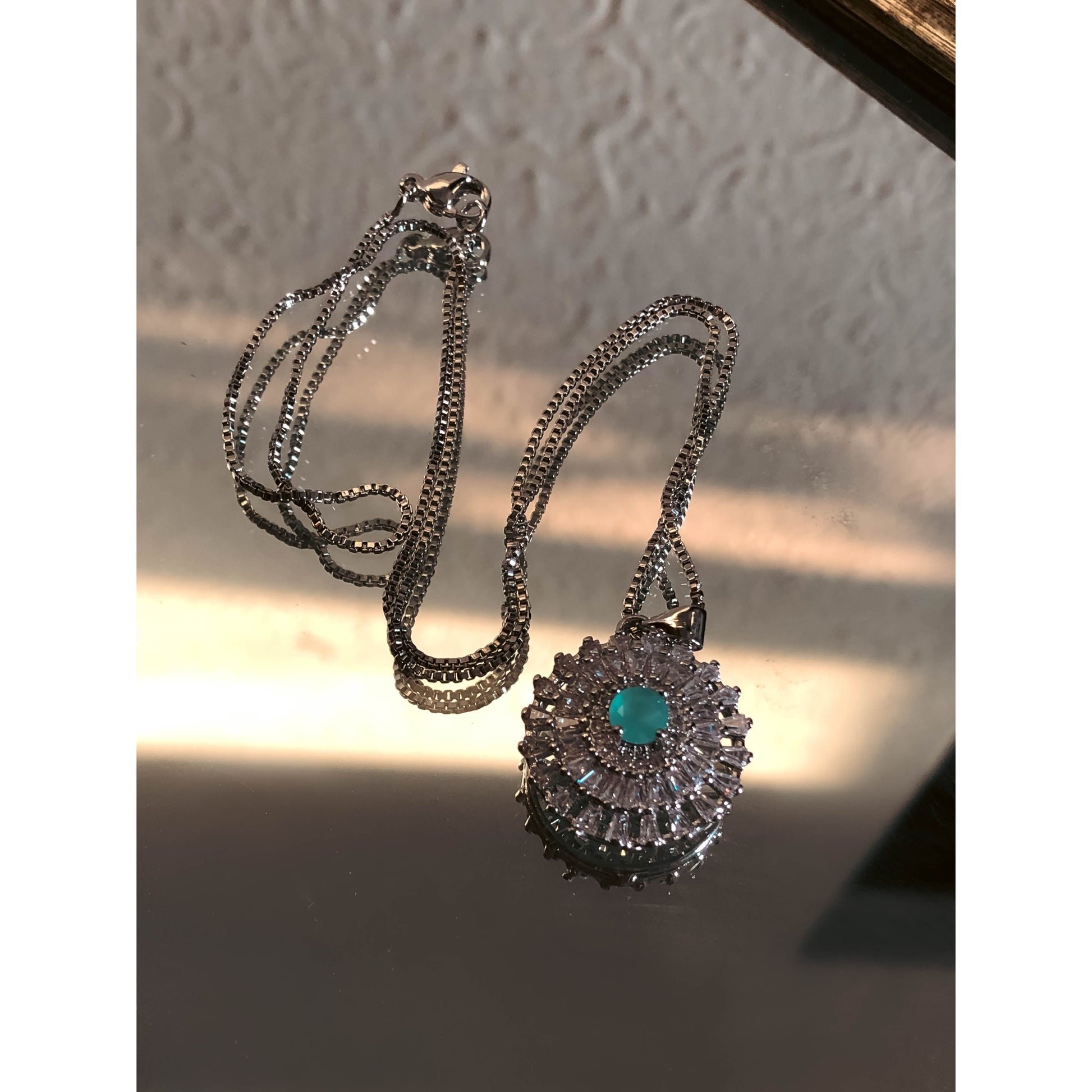 A close-up of a silver-tone necklace with a pendant featuring a large, clear, round stone surrounded by a halo of smaller clear sparkling stones, on a delicate chain.