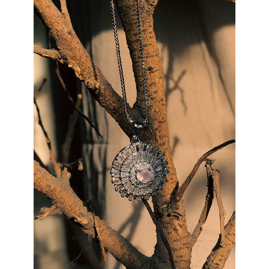 A close-up of a silver-tone necklace with a pendant featuring a large, clear, round stone surrounded by a halo of smaller clear sparkling stones, on a delicate chain.