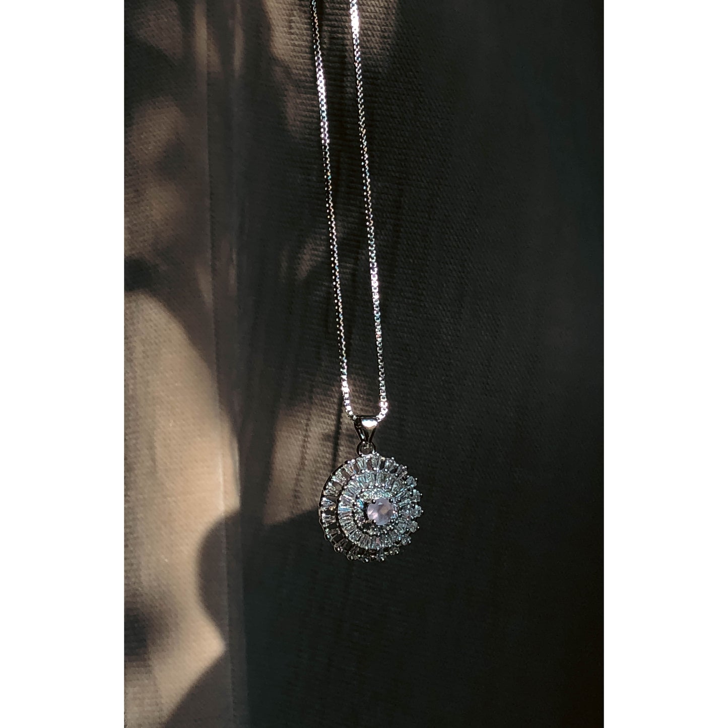 A close-up of a silver-tone necklace with a pendant featuring a large, clear, round stone surrounded by a halo of smaller clear sparkling stones, on a delicate chain.