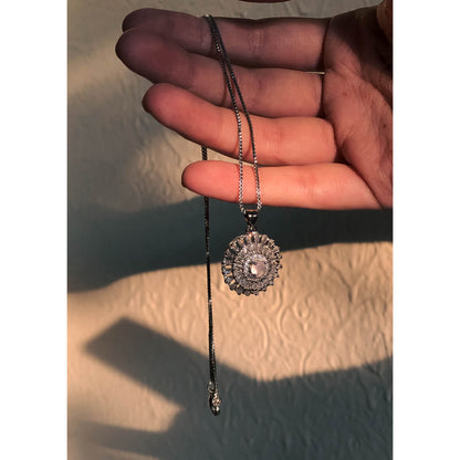 A close-up of a silver-tone necklace with a pendant featuring a large, clear, round stone surrounded by a halo of smaller clear sparkling stones, on a delicate chain.
