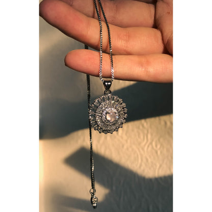 A close-up of a silver-tone necklace with a pendant featuring a large, clear, round stone surrounded by a halo of smaller clear sparkling stones, on a delicate chain.