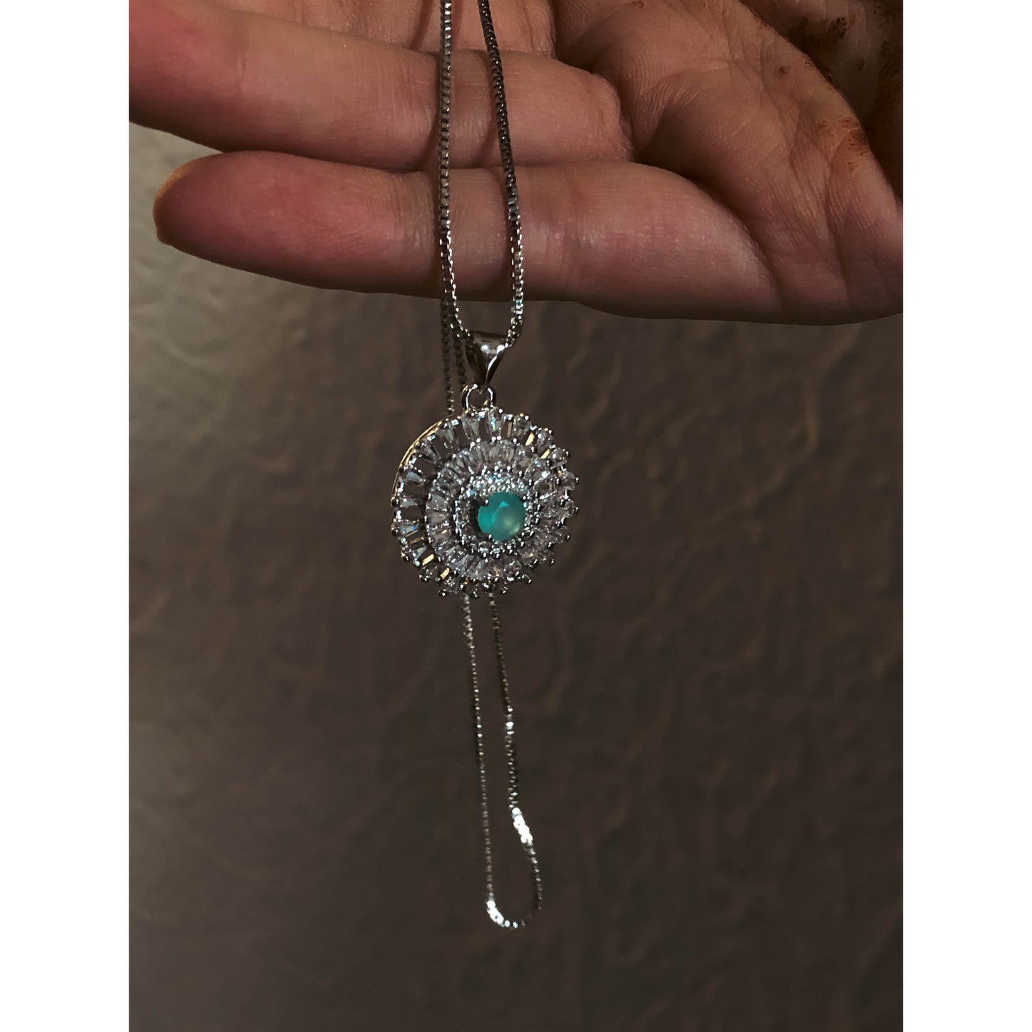 A close-up of a silver-tone necklace with a pendant featuring a large, clear, round stone surrounded by a halo of smaller clear sparkling stones, on a delicate chain.