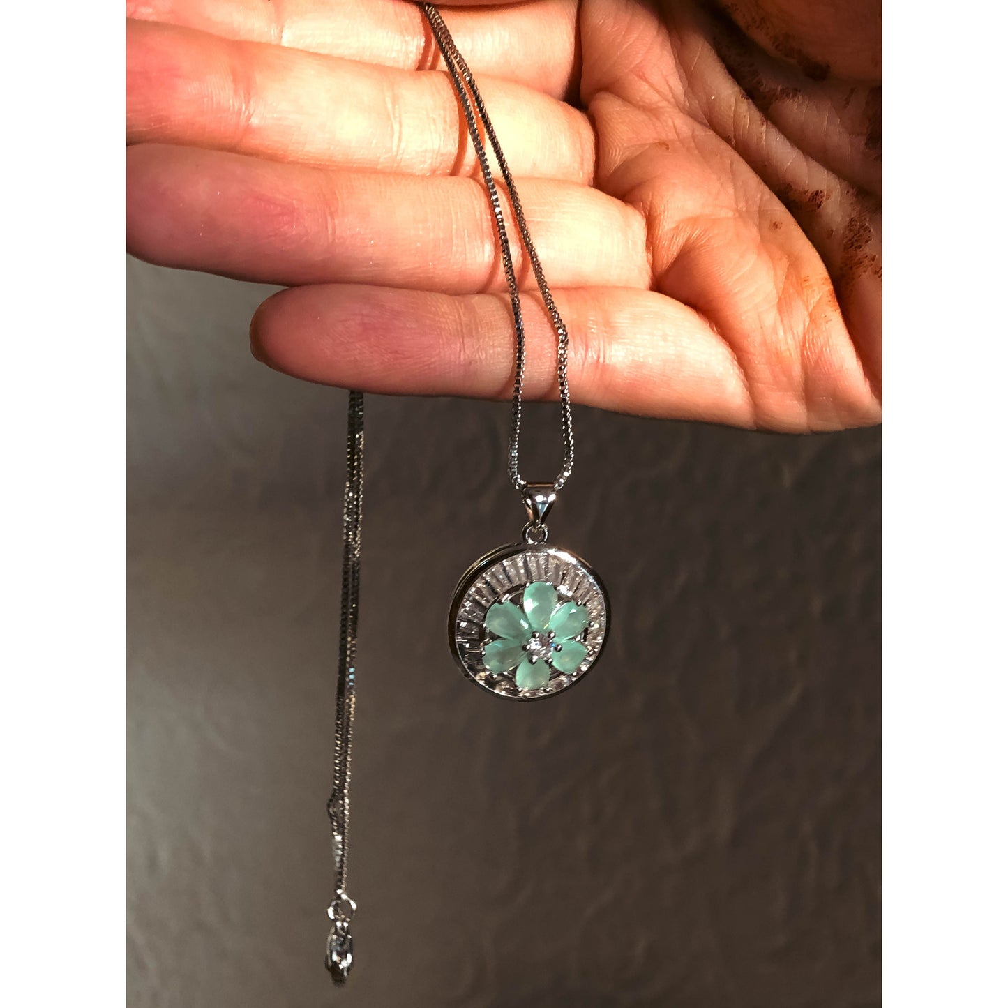 A close-up of a silver-tone necklace with a pendant featuring a clear, flower-shaped cubic zirconia stone surrounded by a halo of smaller clear sparkling stones, on a delicate chain.