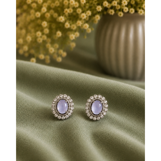 A pair of oval stud earrings with pale lavender center stones surrounded by small clear stones in a silver-toned setting, displayed on draped olive green fabric with blurred yellow flowers in the background.