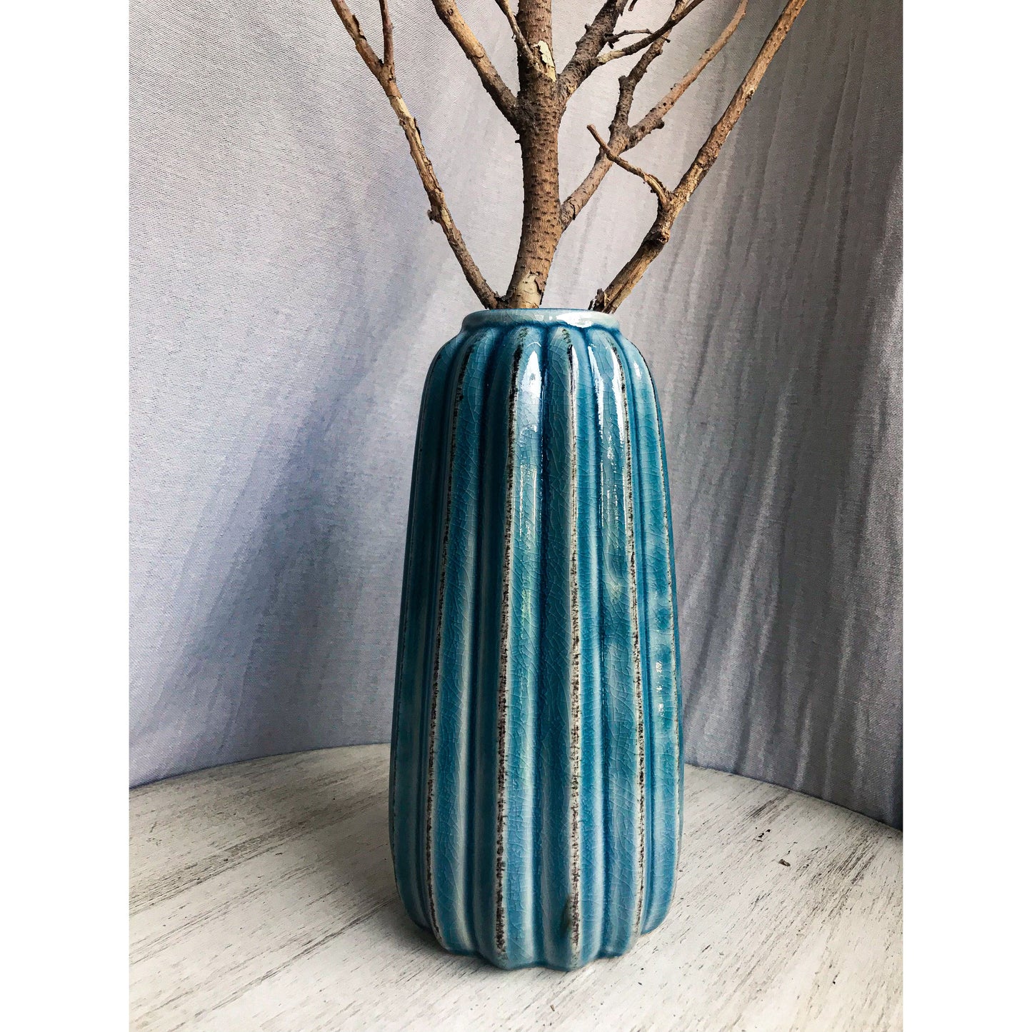 A tall, teal ceramic vase with vertical fluting and a crackled glaze, shown with and without dried branches inside.
