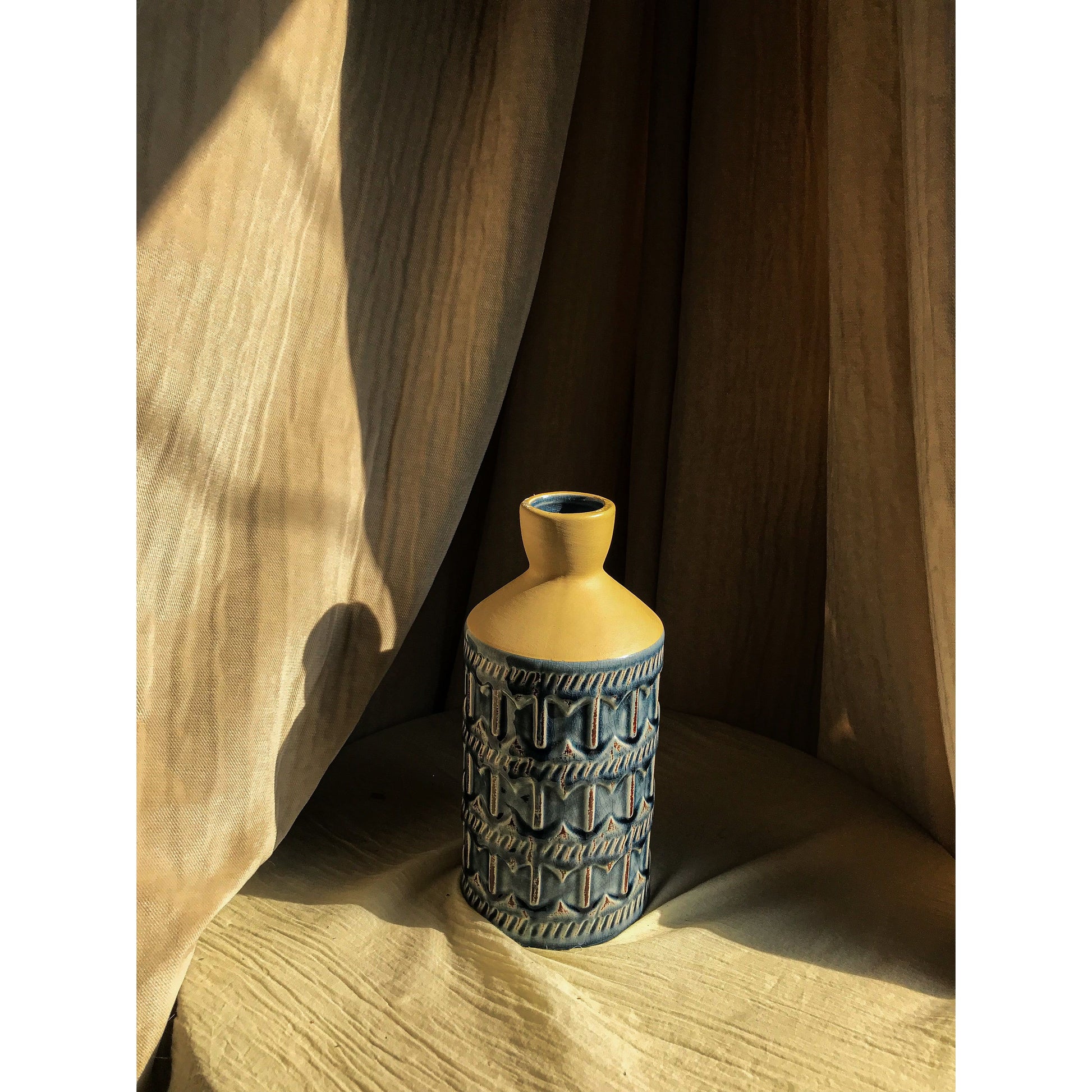 A two-tone ceramic vase with a beige top and a blue bottom featuring an embossed geometric pattern, shown empty and with dried branches.