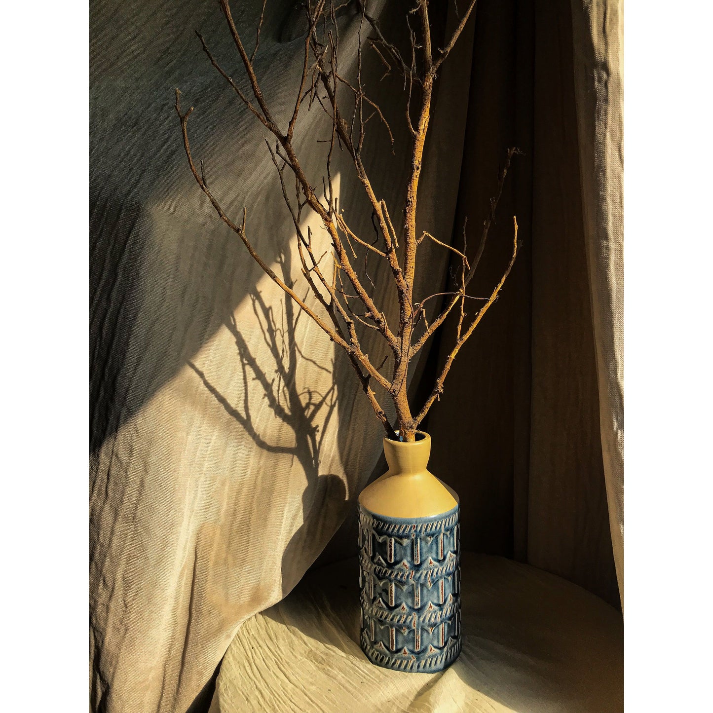A two-tone ceramic vase with a beige top and a blue bottom featuring an embossed geometric pattern, shown empty and with dried branches.
