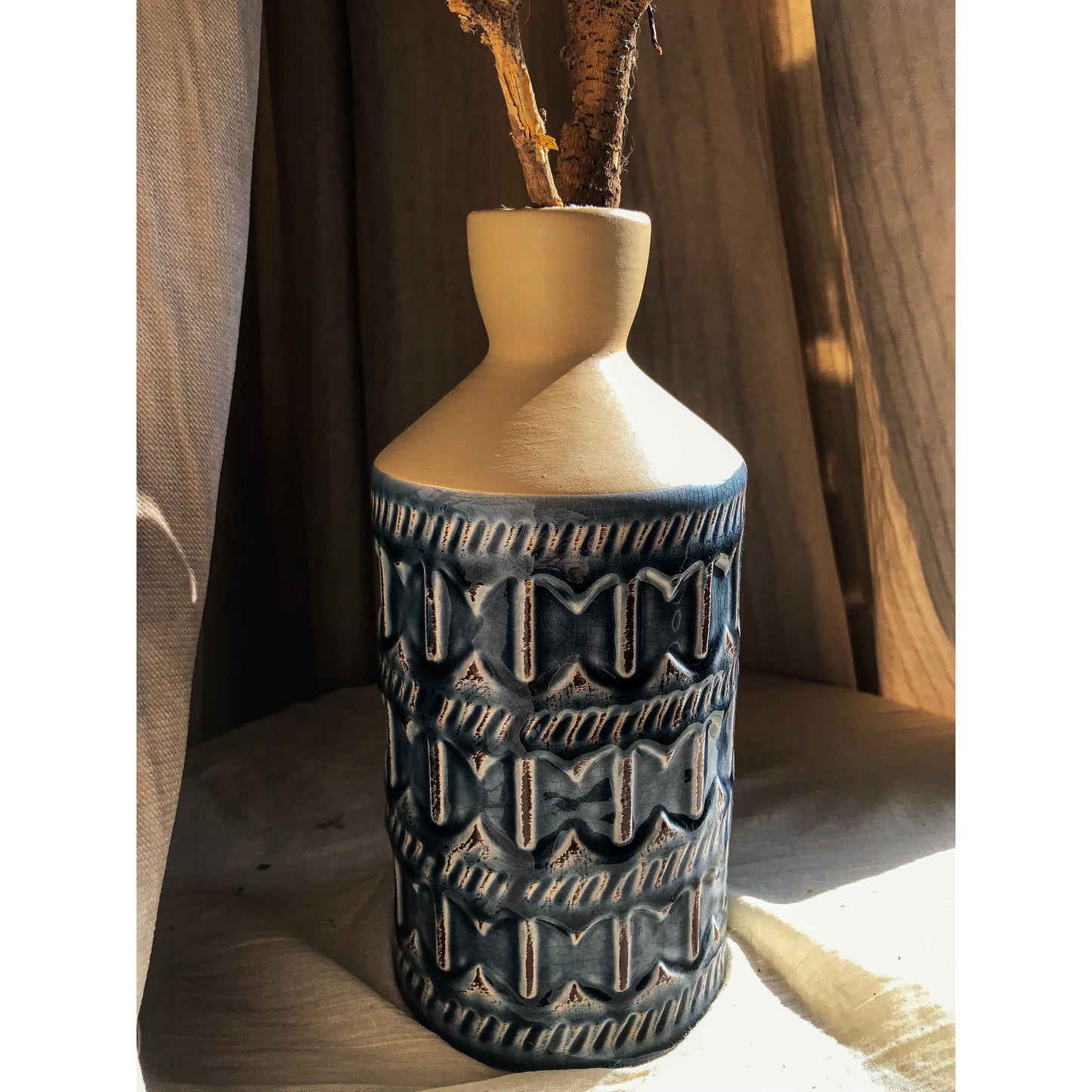 A two-tone ceramic vase with a beige top and a blue bottom featuring an embossed geometric pattern, shown empty and with dried branches.