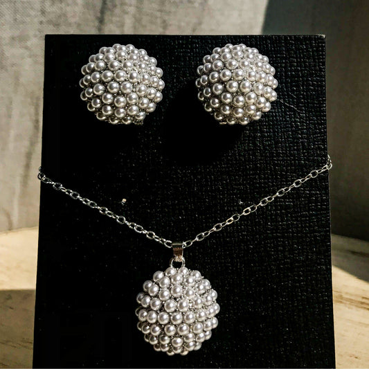 A jewelry set featuring a silver necklace with a round pendant made of many small grey pearls, and a pair of matching round stud earrings made of the same grey pearls, displayed on a black earring card.