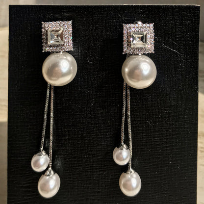 Two silver drop earrings, each with a square top of sparkling clear stones above a large white pearl, from which two chains with smaller white pearls dangle, displayed on a black textured surface.
