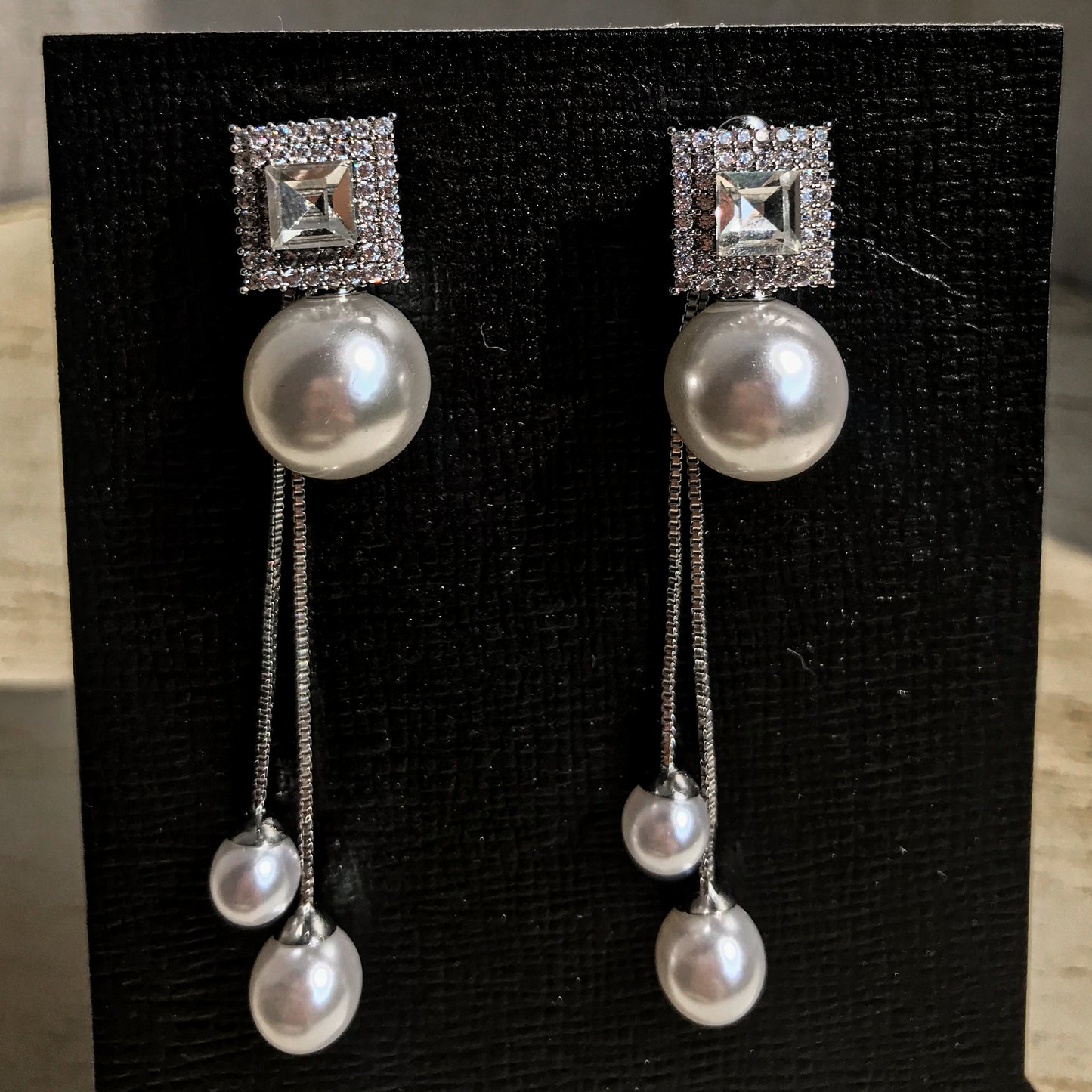 Two silver drop earrings, each with a square top of sparkling clear stones above a large white pearl, from which two chains with smaller white pearls dangle, displayed on a black textured surface.