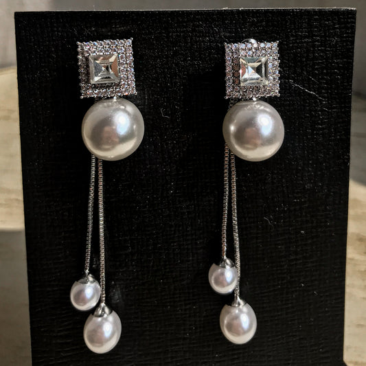 Two silver drop earrings, each with a square top of sparkling clear stones above a large white pearl, from which two chains with smaller white pearls dangle, displayed on a black textured surface.
