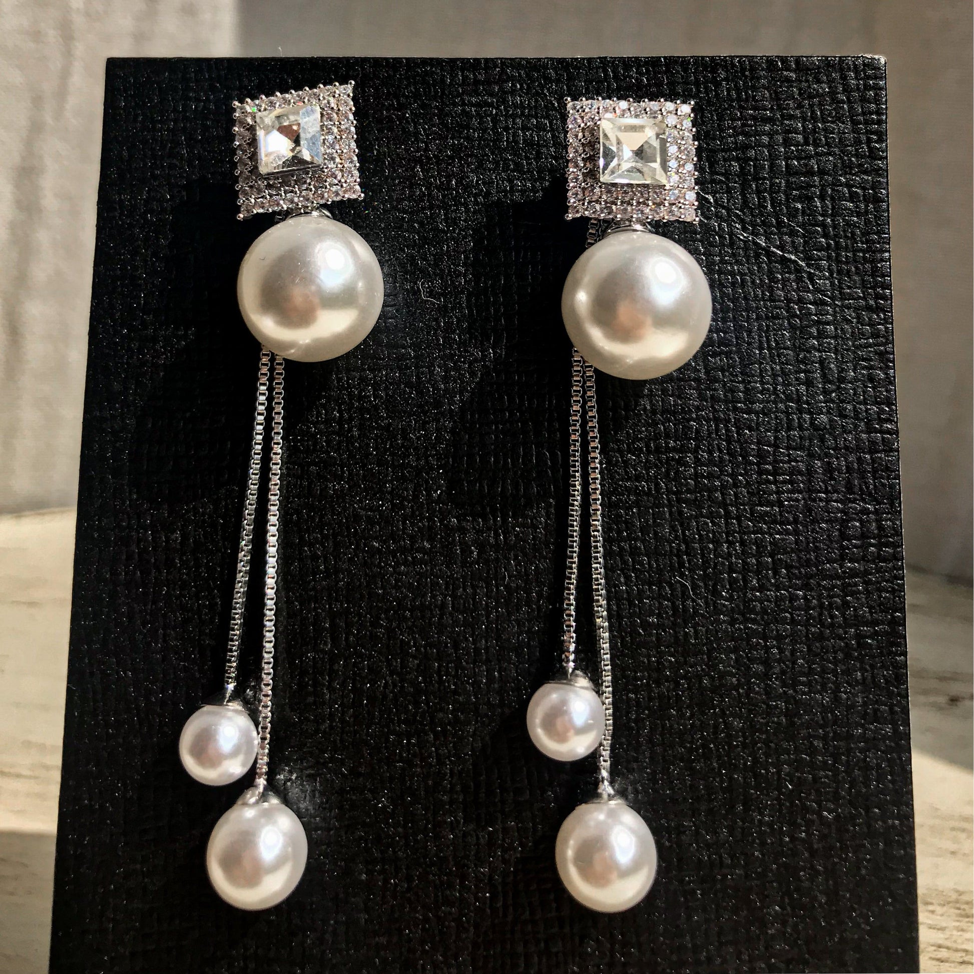 Two silver drop earrings, each with a square top of sparkling clear stones above a large white pearl, from which two chains with smaller white pearls dangle, displayed on a black textured surface.
