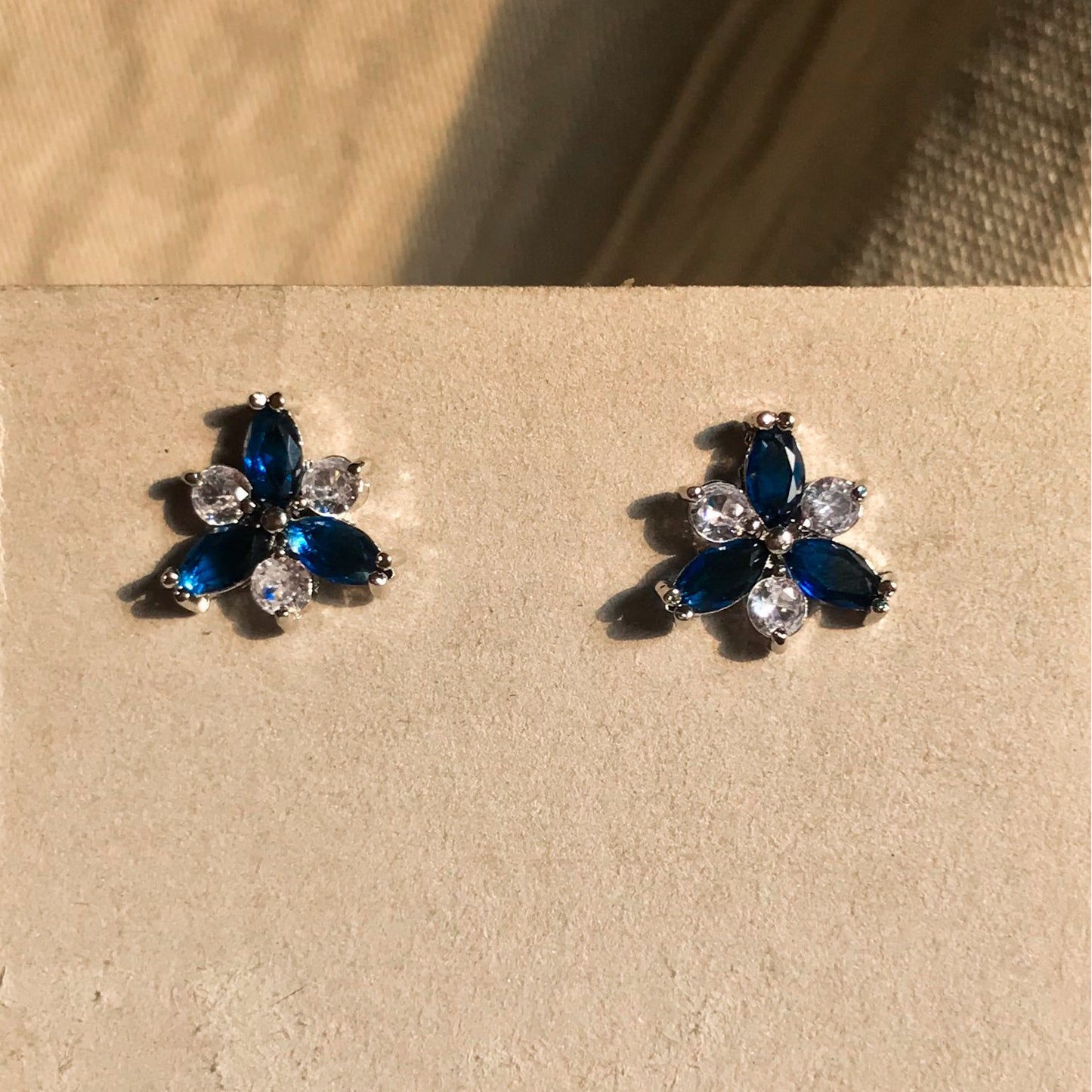 Two small, silver flower-shaped stud earrings set with clear, sparkling stones, displayed on a black earring card.