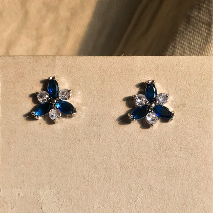 Two small, silver flower-shaped stud earrings set with clear, sparkling stones, displayed on a black earring card.