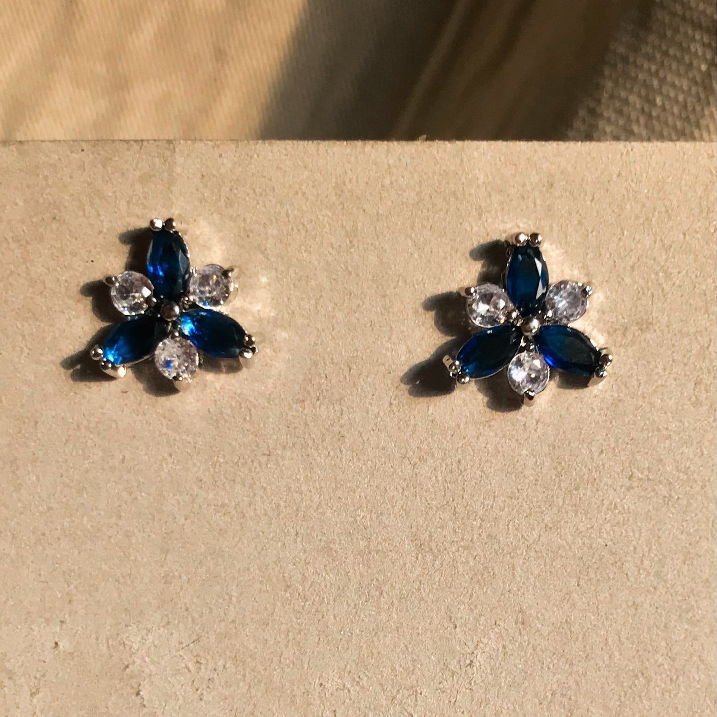 Two small, silver flower-shaped stud earrings set with clear, sparkling stones, displayed on a black earring card.