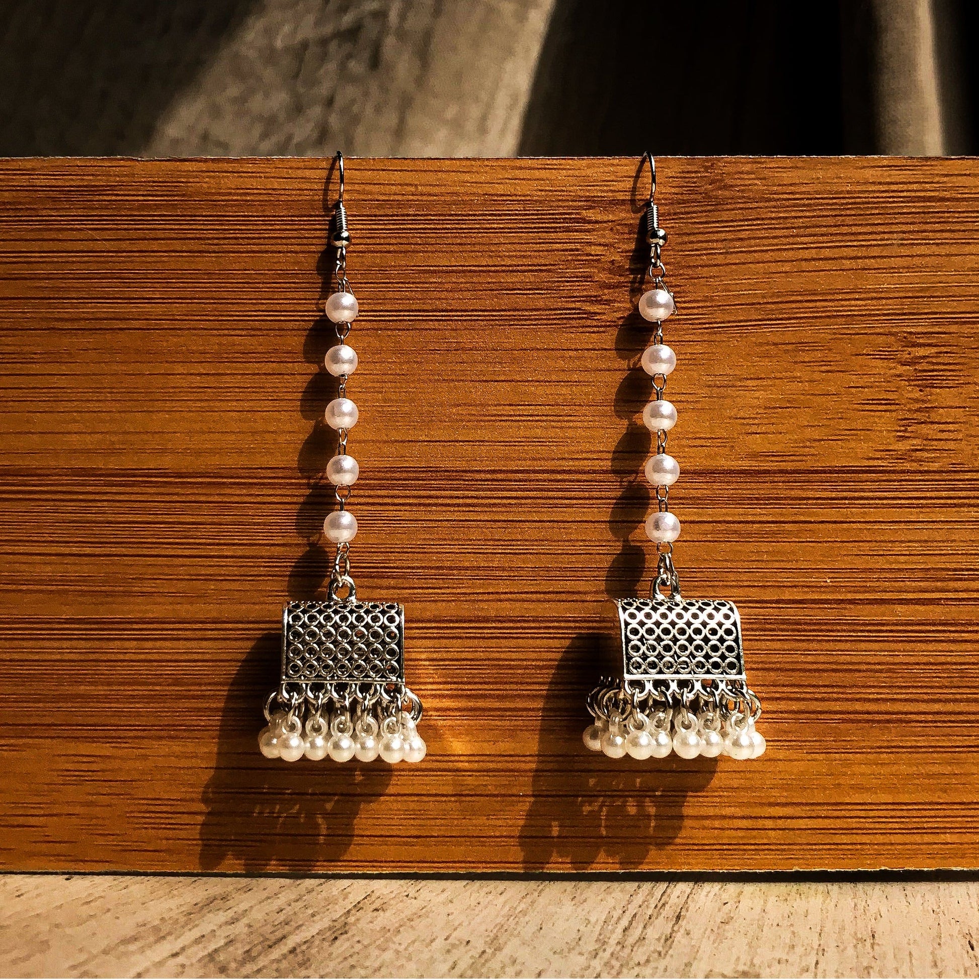 Two silver-tone dangle earrings with a textured geometric top, a chain of white pearls, and a fringe of small white pearls at the bottom, displayed against a wooden background.