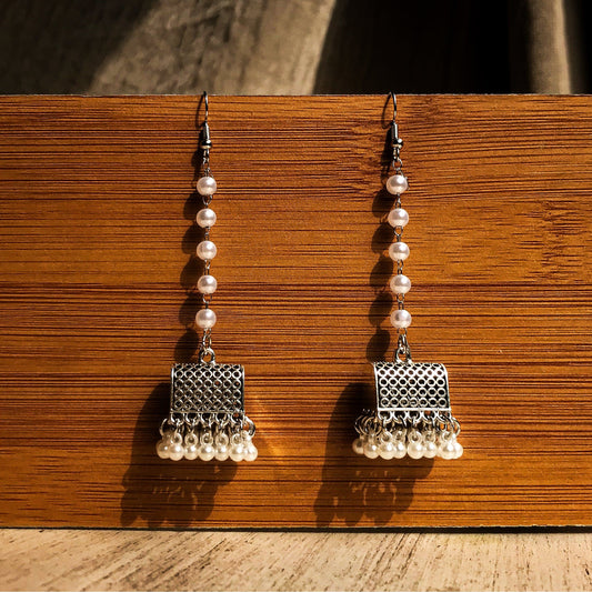 Two silver-tone dangle earrings with a textured geometric top, a chain of white pearls, and a fringe of small white pearls at the bottom, displayed against a wooden background.