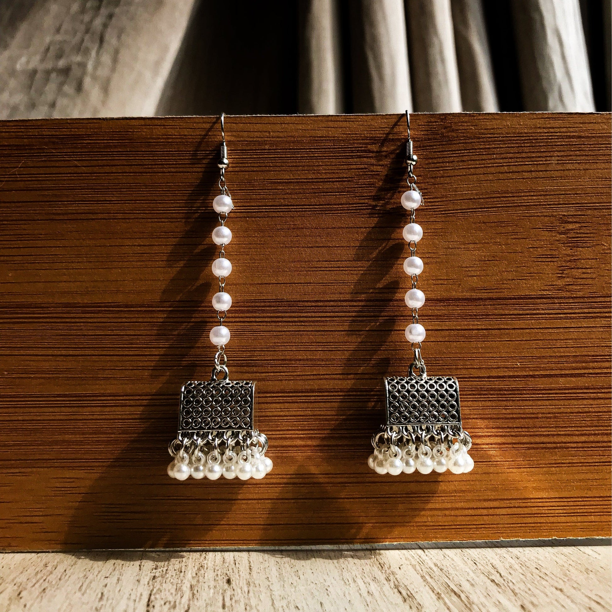 Two silver-tone dangle earrings with a textured geometric top, a chain of white pearls, and a fringe of small white pearls at the bottom, displayed against a wooden background.
