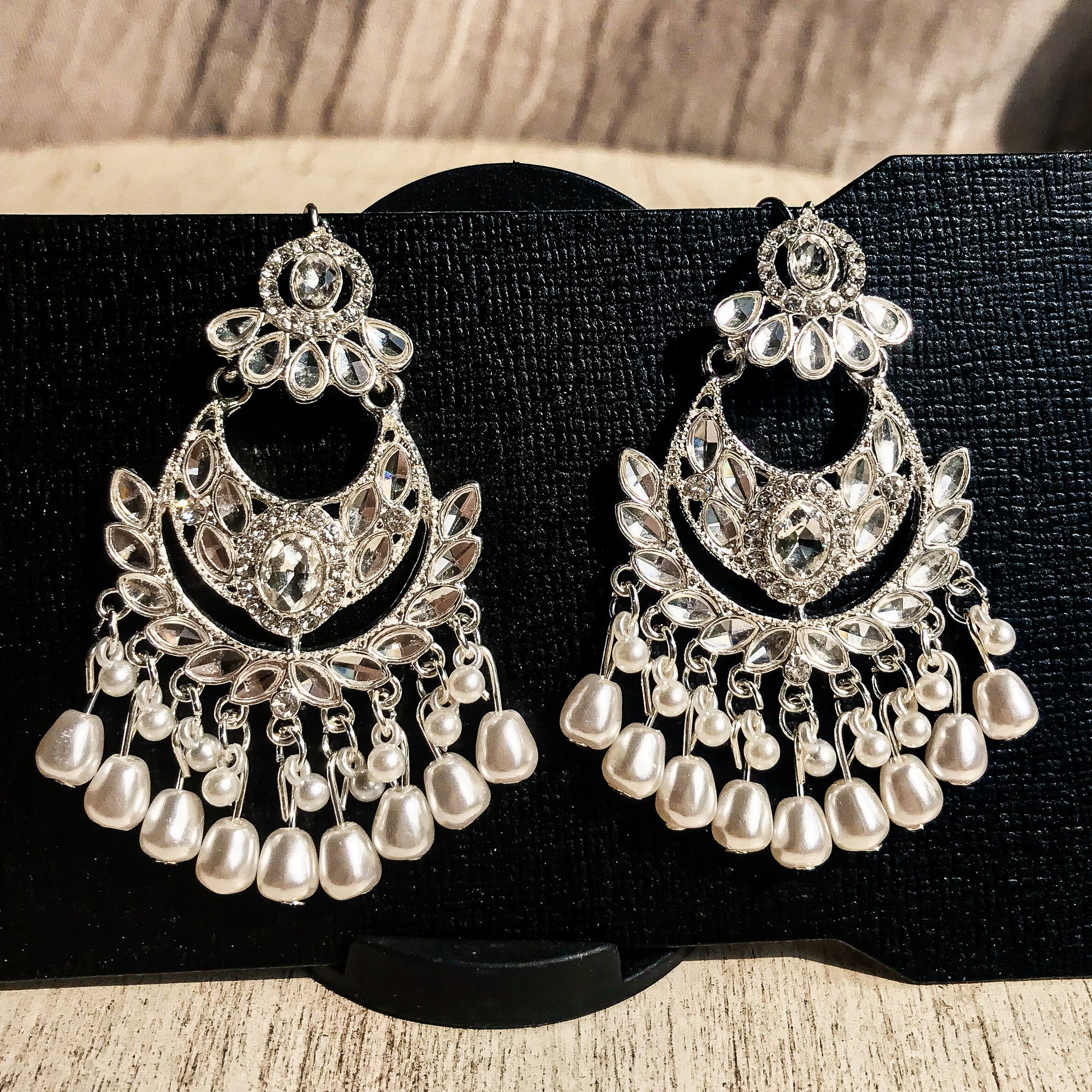 Two ornate silver-tone chandelier earrings with multiple clear crystals and cascading strands of white pearls, displayed on a black earring card.