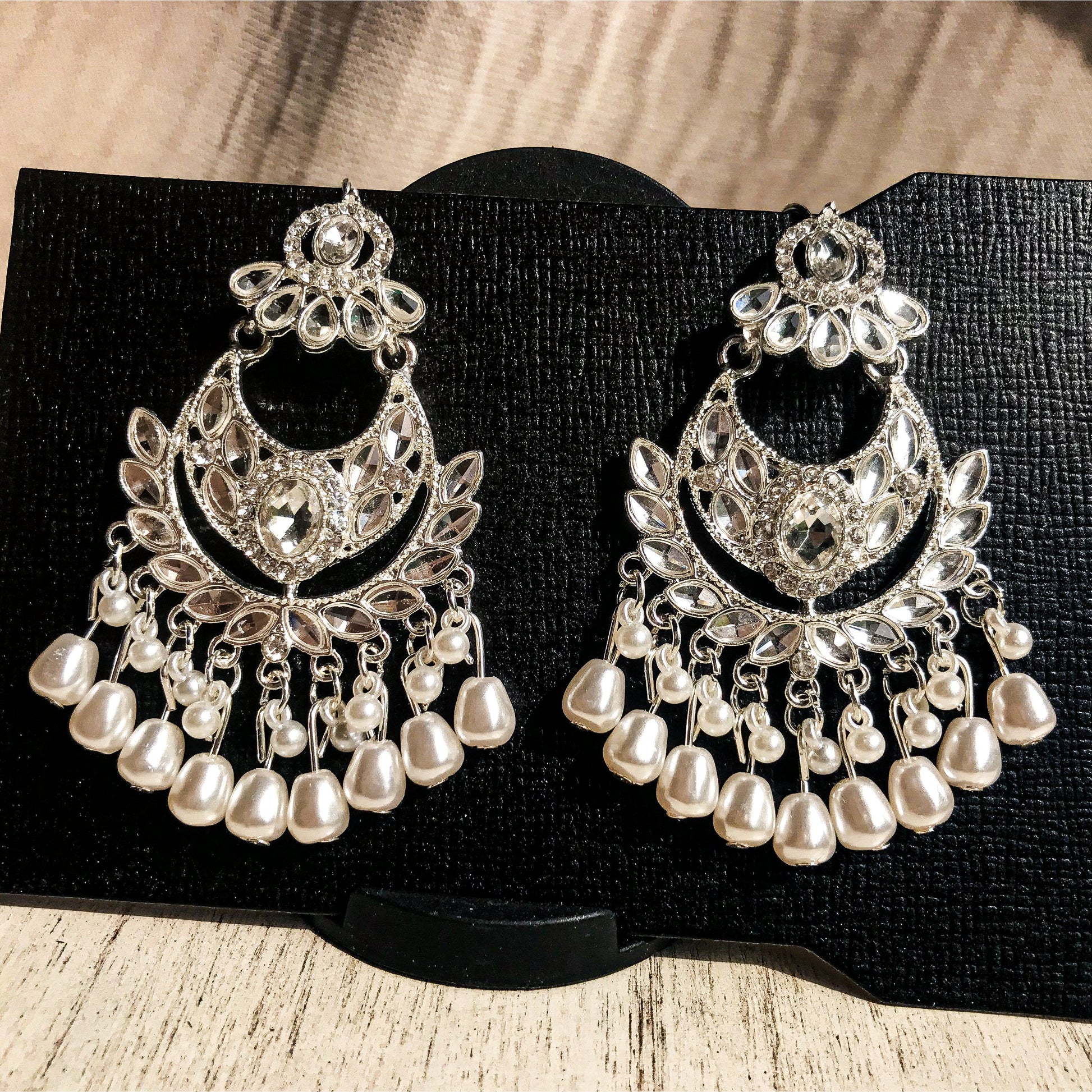 Two ornate silver-tone chandelier earrings with multiple clear crystals and cascading strands of white pearls, displayed on a black earring card.