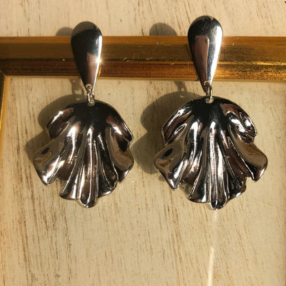 Two sculptural metal drop earrings, one silver and one gold, with a flowing, draped design, displayed against a framed background.
